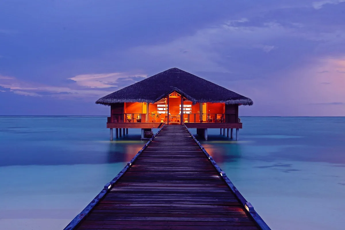 Medhufushi Island Resort, spa, Meemu Atoll, Maldives, Over-water spa pavilion at twilight: symmetrical thatched-roof pavilion at the end of a long timber jetty lined with low LED bollards, warm orange-lit interior glowing through floor-to-ceiling glass walls, deep purple-and-pink twilight sky reflecting on the perfectly calm lagoon, the property's signature spa composition at the dusk hour.