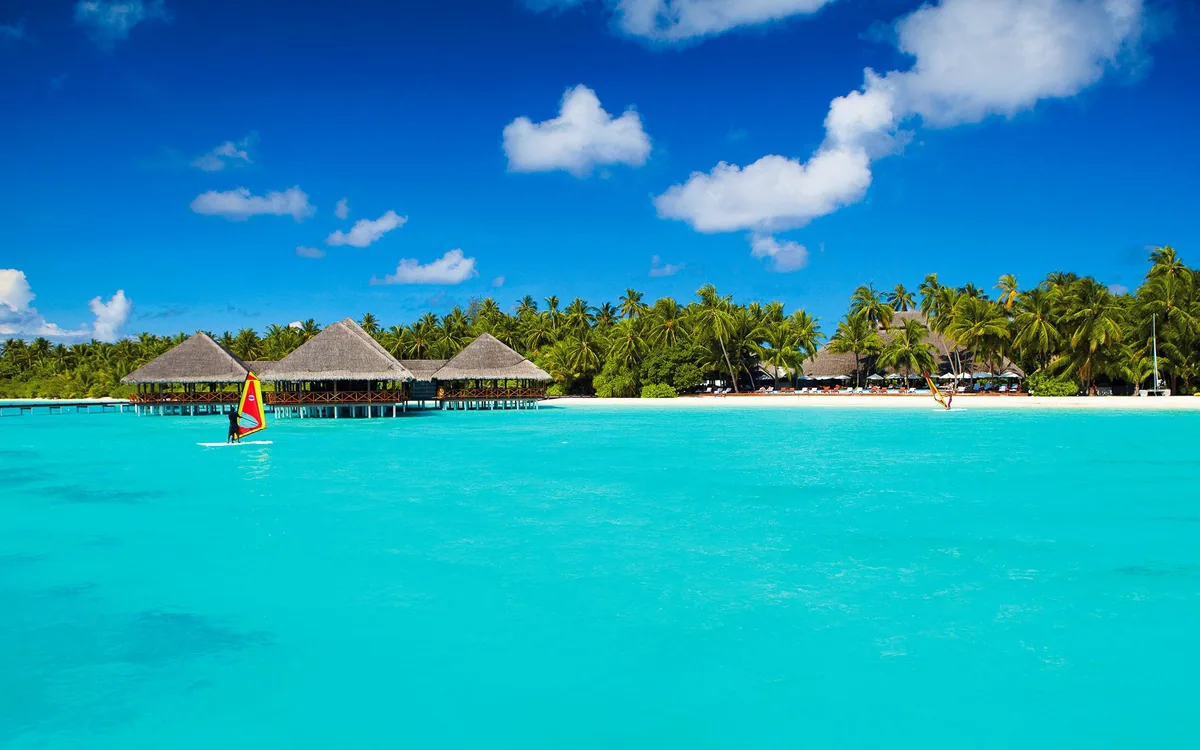 Medhufushi Island Resort, setting, Meemu Atoll, Maldives, Lagoon view toward the island: the distinctive cluster of three thatched conical-roof overwater pavilions on stilts (the dining cluster), a small windsurfer with a red-and-yellow sail in the foreground lagoon water, the palm-covered island stretching to the right with more thatched roofs glimpsed behind the trees, vibrant turquoise water, a blue sky scattered with clouds.