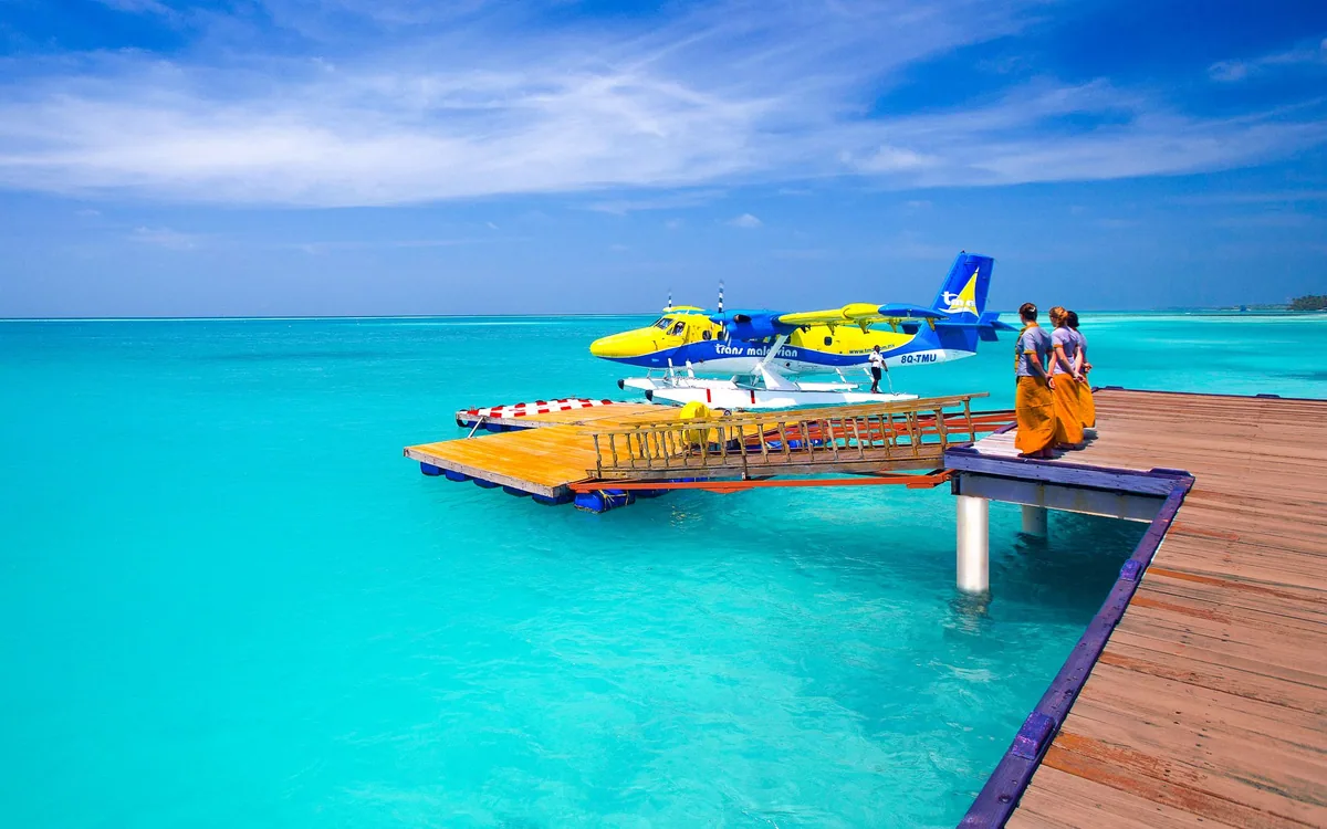 Medhufushi Island Resort, lifestyle, Meemu Atoll, Maldives, Seaplane arrival: a Trans Maldivian Airways yellow-and-blue Twin Otter (with the operator branding visible) docked at the property's wooden arrival jetty, a guest couple greeted by staff in traditional orange Maldivian sarong, the vibrant turquoise lagoon framing the composition, a blue sky scattered with clouds overhead.