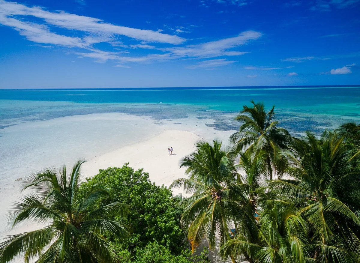 Medhufushi Island Resort, lifestyle, Meemu Atoll, Maldives, Beach-walk aerial: a couple walking on the white sand beach captured from the high palm canopy looking down, the property's signature elongated sand-tip composition extending into the lagoon, dense coconut palms framing the perimeter, vibrant turquoise water meeting the open ocean at the horizon line.