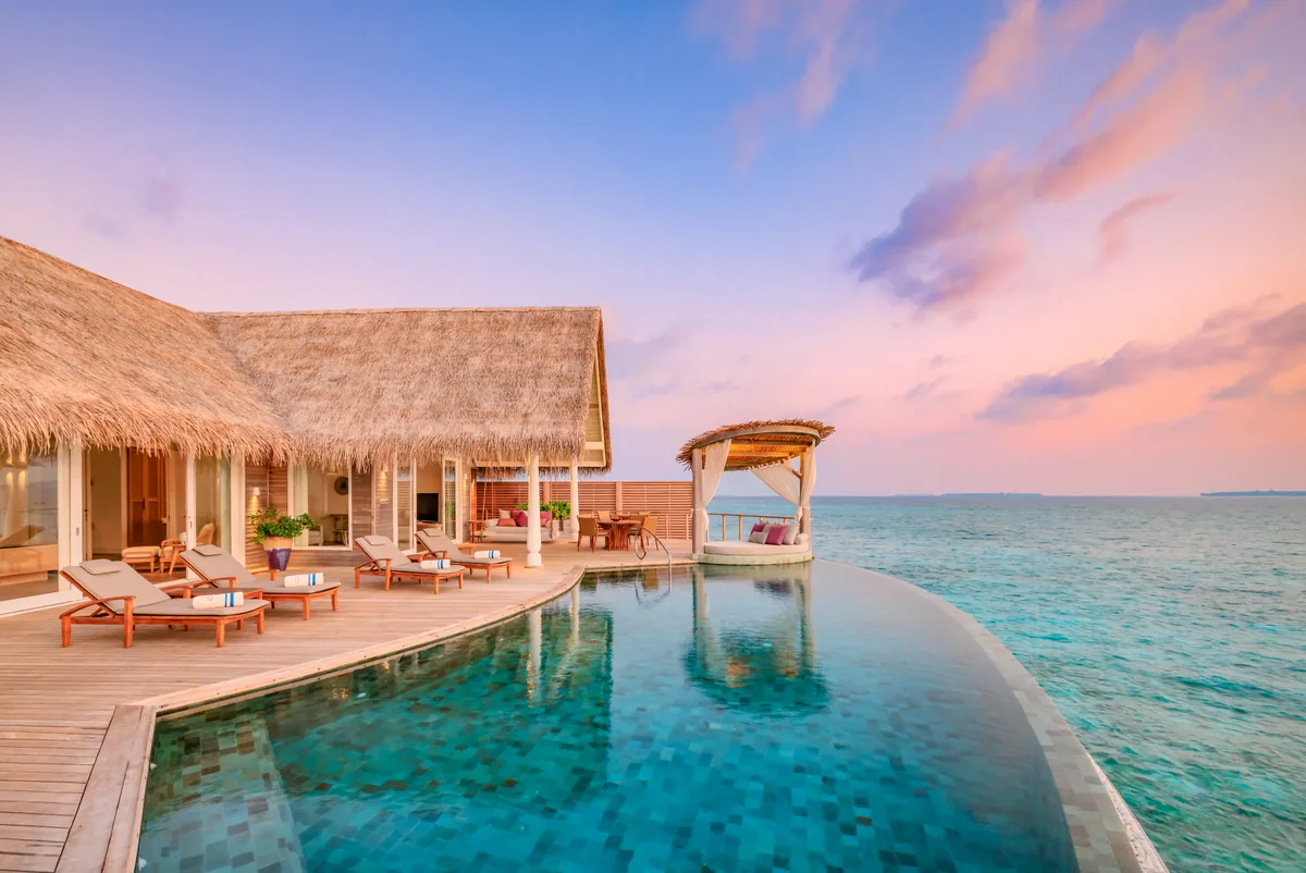 Milaidhoo Island Maldives, villa overwater, Baa Atoll, Maldives, Ocean Residence private deck at twilight, the long mosaic-tile infinity pool meeting the lagoon edge, five cream-cushioned sun-loungers in formation, freestanding canopy daybed with pink cushion and the peach-pink twilight reflecting on the still water.
