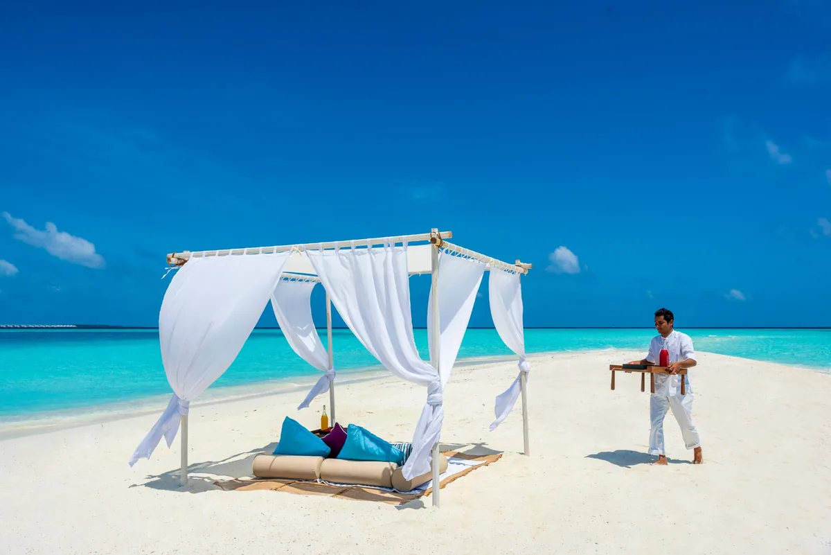 Milaidhoo Island Maldives, dining, Baa Atoll, Maldives, Private sandbank destination dining setup, the white-curtained four-poster cabana with cream-cushion daybed and teal-and-magenta pillows on the sun-bleached sand, a white-uniformed server carrying the cobalt-blue welcome-drink bottle on a timber tray.