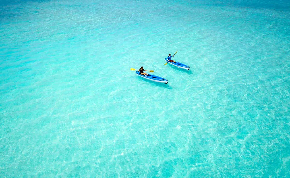 Milaidhoo Island Maldives, activities, Baa Atoll, Maldives, Two single kayaks drifting through the glass-clear lagoon shallows from the drone-height view, the pale-turquoise water revealing the sandy floor in clean detail and the two paddlers in casual swimwear navigating in formation.