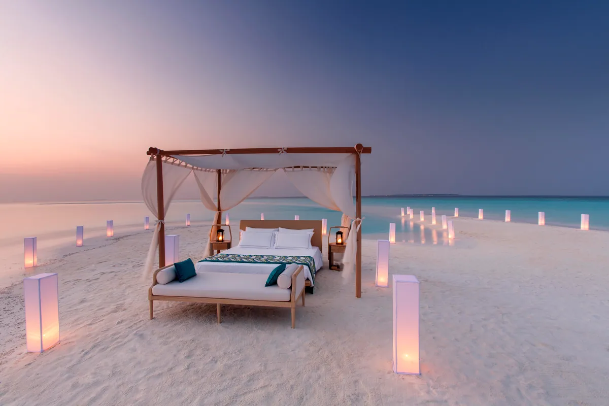 Milaidhoo Island Maldives, activities, Baa Atoll, Maldives, Sleeping Under the Stars sandbank setup at twilight, a four-poster timber bed with cream sheer curtains and white-pillow bedding on the sun-bleached sandbank, brass storm-lanterns flanking the bed, twin runways of pink-paper-lantern luminaria leading out toward the lagoon and the lavender-and-blue dusk sky stretching to the horizon.