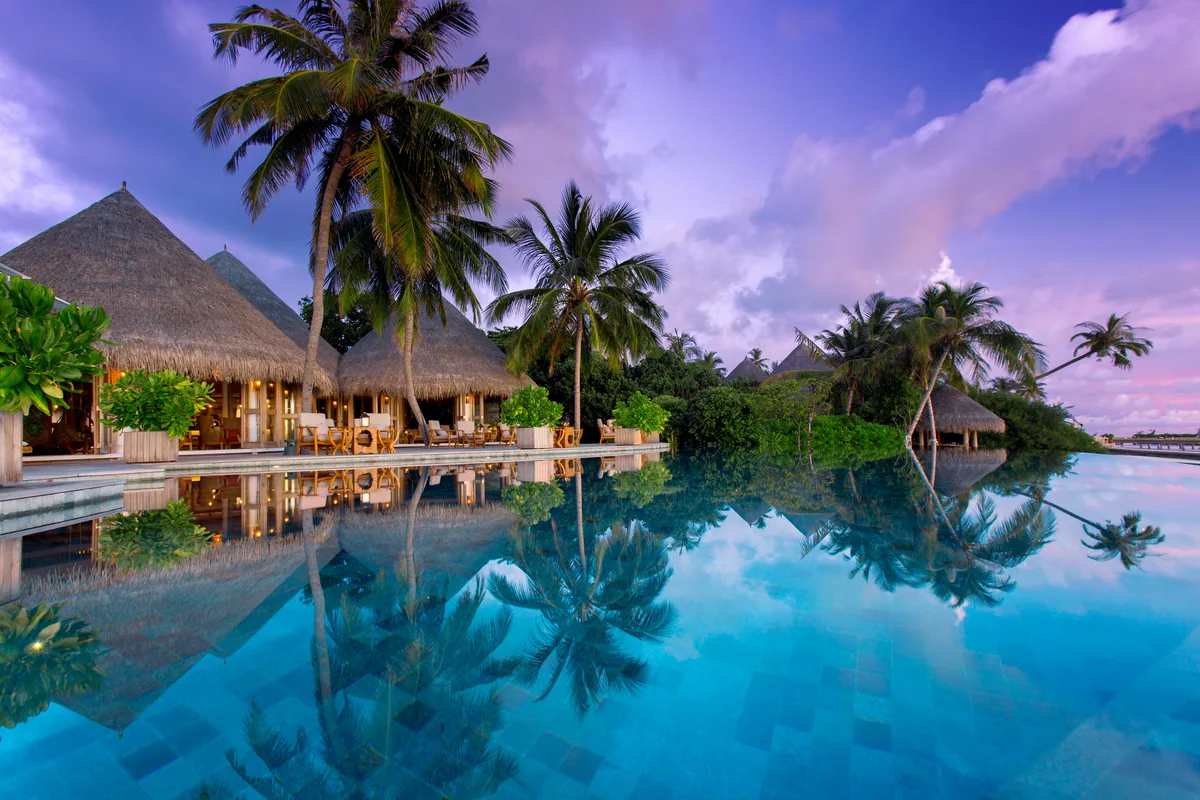 Milaidhoo Island Maldives, dining, Baa Atoll, Maldives, Compass Pool Bar at twilight, the thatched-roof bar pavilion glowing warm beside the infinity-edge mosaic-tile pool, palm silhouettes reflected on the glass-still water and the lavender-and-pink sunset sky stretching to the horizon.
