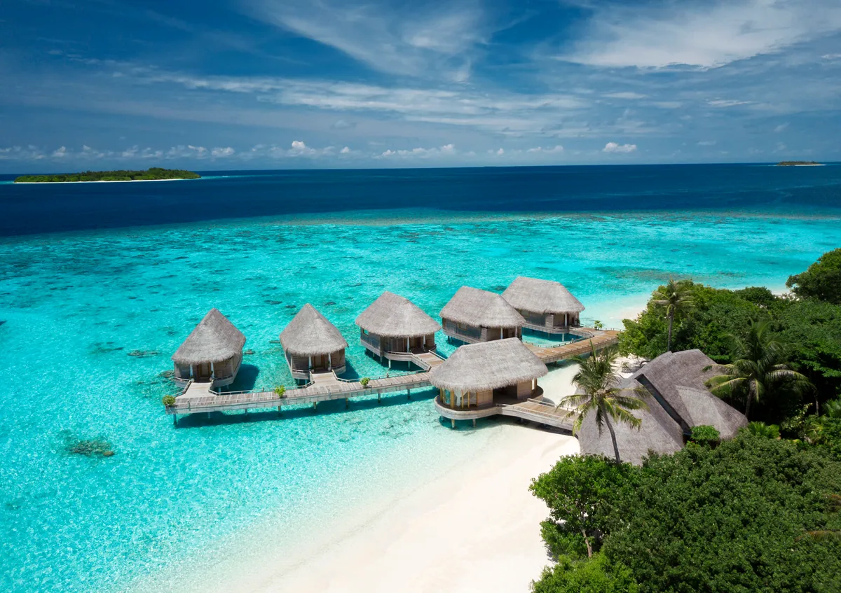 Milaidhoo Island Maldives, spa, Baa Atoll, Maldives, Serenity Spa from the drone height, the four thatched-roof overwater treatment suites in formation off the white-sand beach, the larger thatched reception pavilion at the beach connection and the surrounding reef shelf in shallow turquoise water.