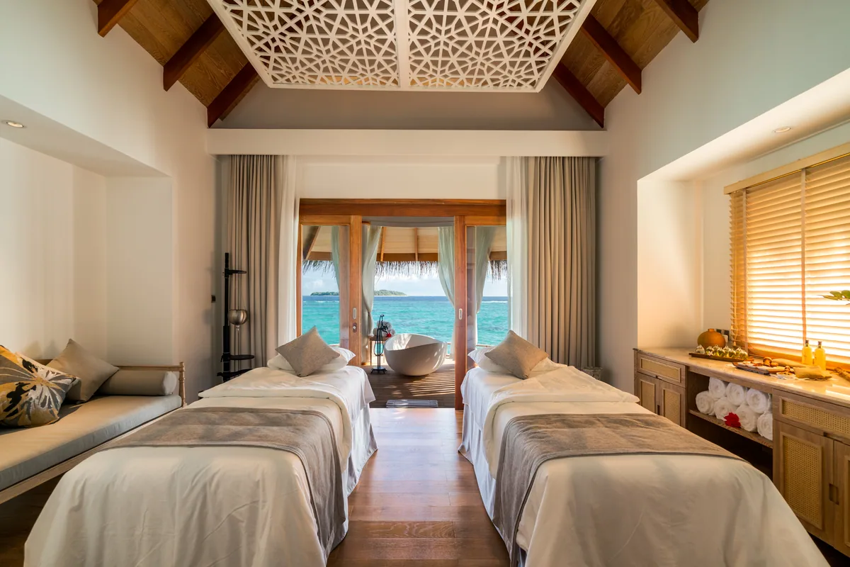 Milaidhoo Island Maldives, spa, Baa Atoll, Maldives, Serenity Spa twin treatment-suite interior, the peaked timber-rafter ceiling with the carved geometric panel above the paired massage beds, freestanding white sculpted bathtub framed by cream curtains opening directly to the lagoon and the spa pavilion across the water.