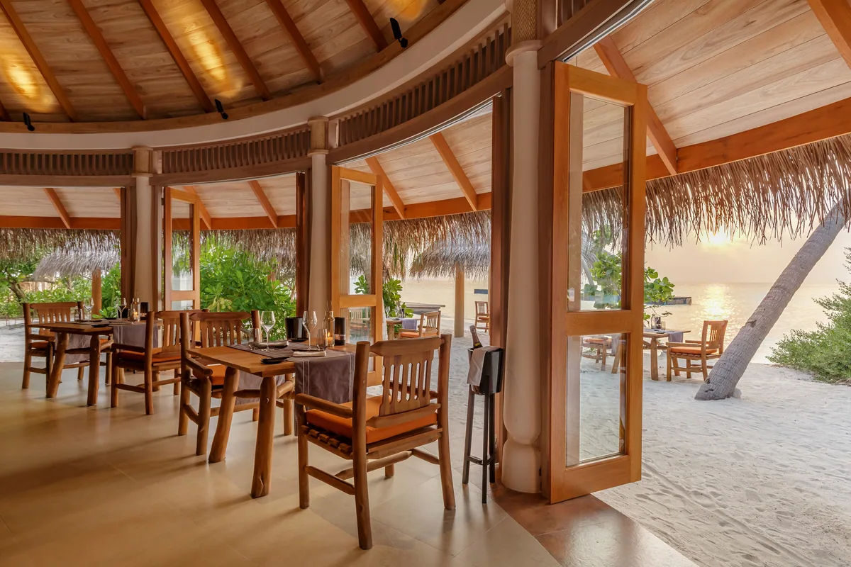 Milaidhoo Island Maldives, dining, Baa Atoll, Maldives, Shoreline Grill interior, the warm timber-rafter ceiling above dark-wood tables with terracotta-orange cushioned chairs, the open eaves with thatched-eyebrow fringe opening directly to the white-sand beach and the lagoon framed by a single coconut palm.