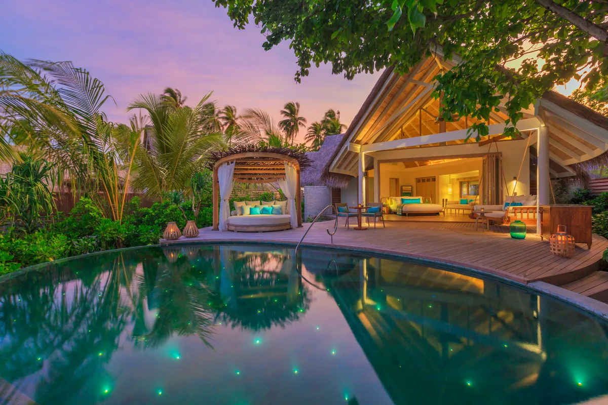 Milaidhoo Island Maldives, villa beach, Baa Atoll, Maldives, Beach Pool Villa at twilight, the freeform plunge pool studded with underwater fibre-optic starlight points, daybed cabana with teal cushions on the timber deck, peaked thatched-roof villa interior glowing warm behind the sliding doors and dense palm-grove framing the lavender sky.