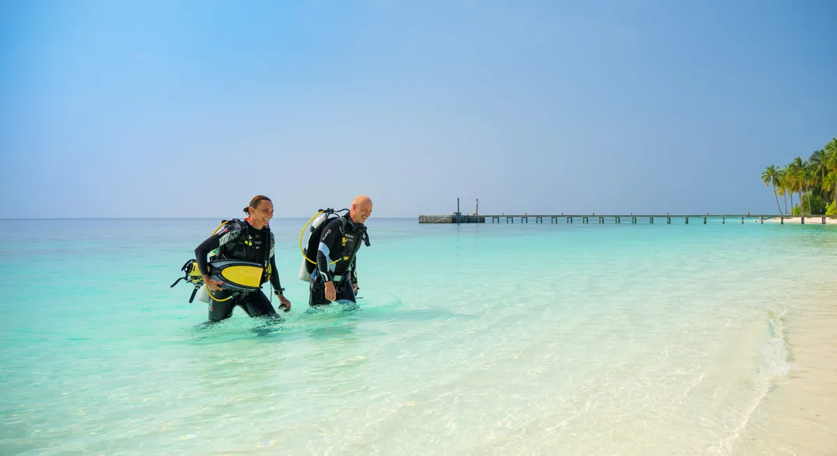 Mirihi Island Resort, activities, Alif Dhaal (South Ari) Atoll, Maldives, Divers in full scuba kit walking out of the lagoon at the western jetty, the daily house-reef shore-exit pattern at the 5-to-15-metre reef-edge distance.