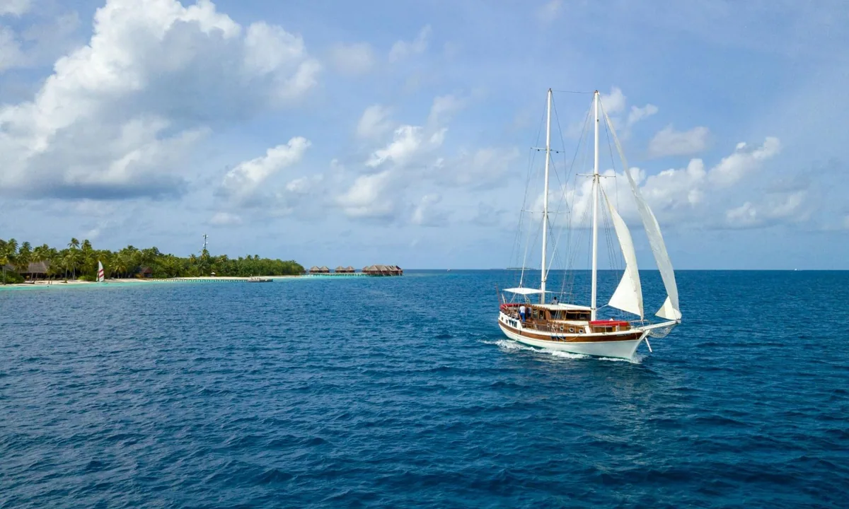 Mirihi Island Resort, activities, Alif Dhaal (South Ari) Atoll, Maldives, Property-branded Thari two-mast wooden sailing schooner under partial sail with Mirihi's eastern jetty cluster visible at the left horizon.