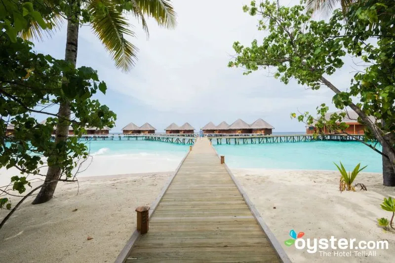 Mirihi Island Resort, setting, Alif Dhaal (South Ari) Atoll, Maldives, Wooden boardwalk leading from the beach to the over-water villa cluster at the reef edge, palm canopy framing the approach and thatch villa silhouettes on the lagoon, the property's primary arrival composition.
