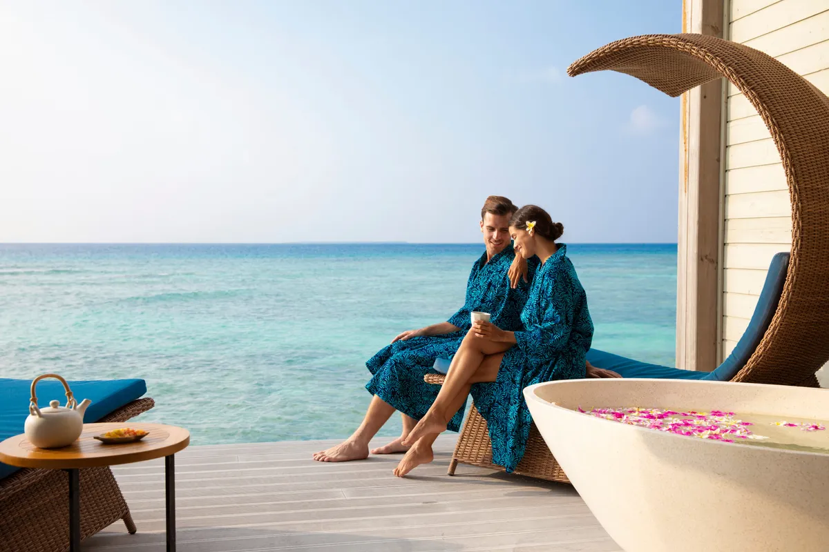Mövenpick Resort Kuredhivaru, Zara Spa over-water pavilion deck with couple in robes beside hibiscus-petal stone bath, Noonu Atoll, Maldives