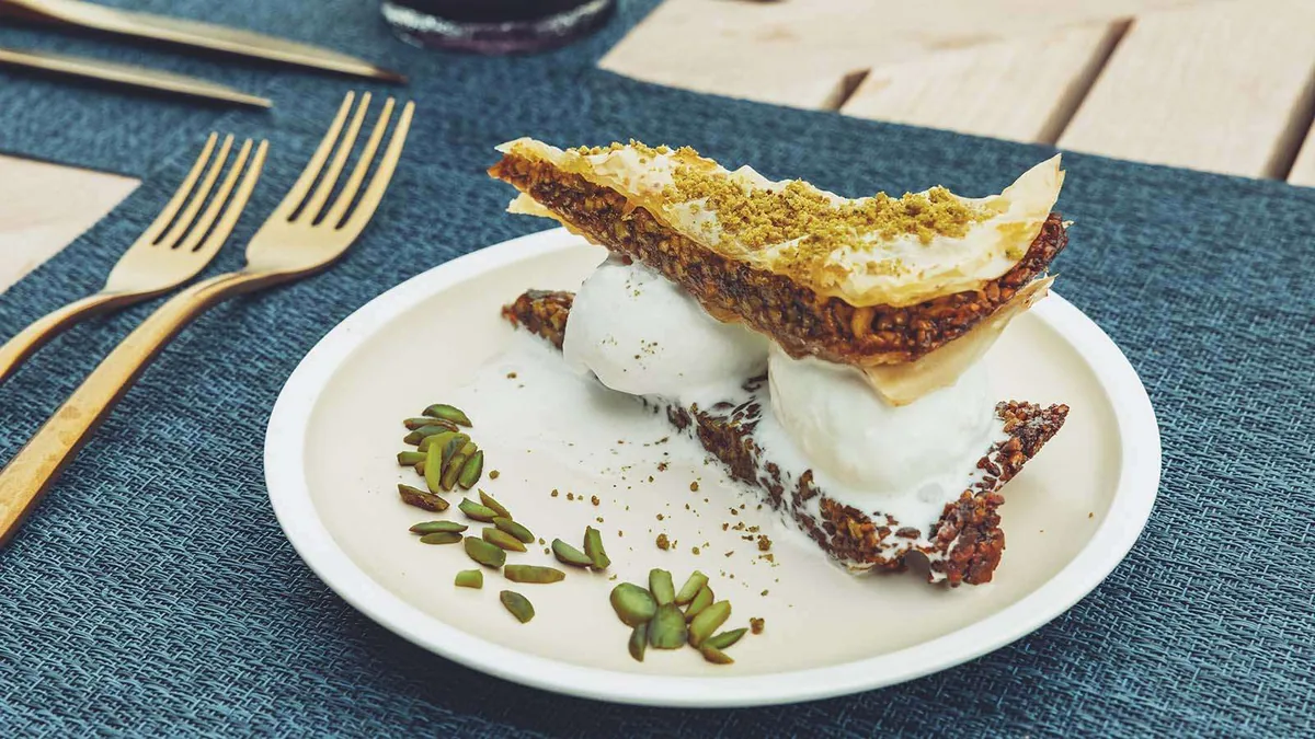 Nala Maldives by Jawakara, dining, Lhaviyani Atoll, Maldives, Milos Greek venue dessert plating: pistachio-and-honey baklava with vanilla ice-cream scoops, pistachio-shell scatter, gold cutlery on a blue placemat.