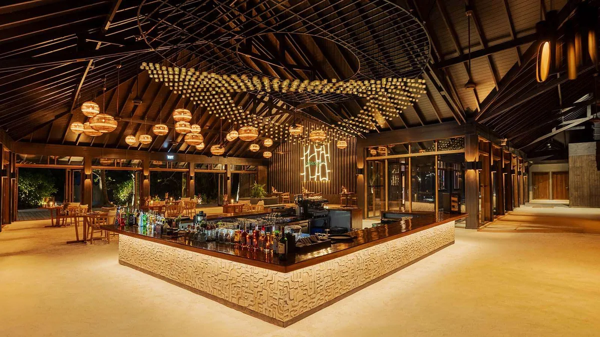 Nala Maldives by Jawakara, dining, Lhaviyani Atoll, Maldives, Velu Bar large open pavilion at night with a hexagonal corner bar carved with tribal pattern fronting, a cascading light-cube ceiling installation, woven globe pendants, and the Velu Bar neon sign on the back wall.