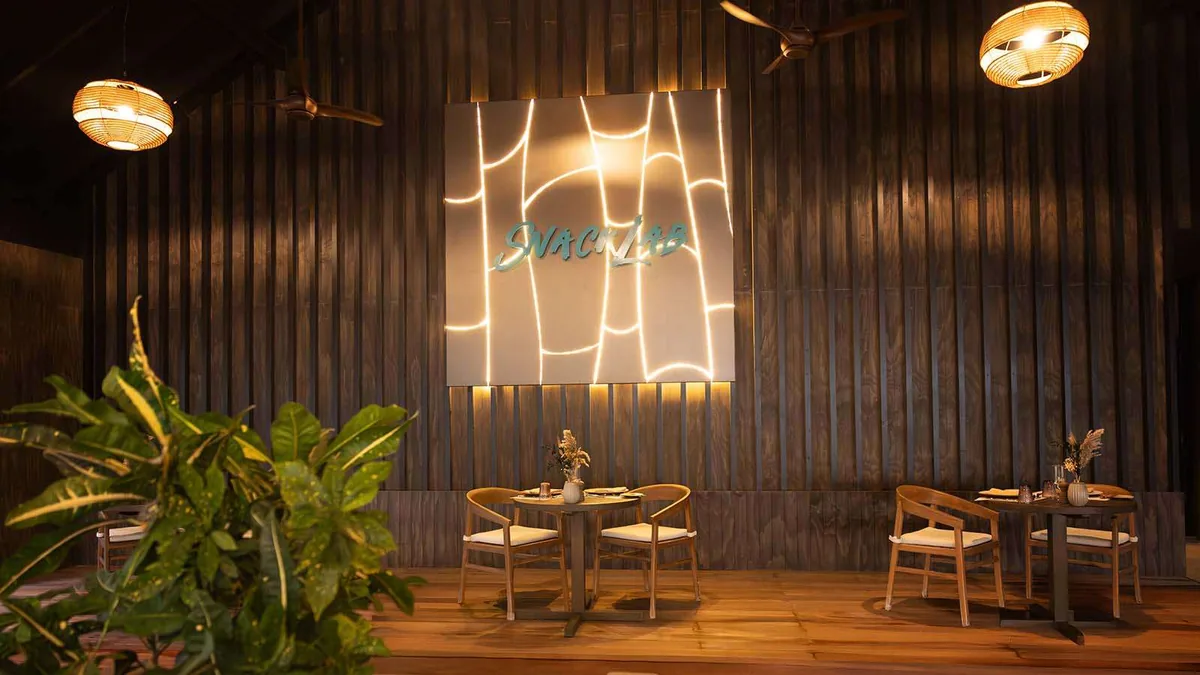 Nala Maldives by Jawakara, dining, Lhaviyani Atoll, Maldives, Snack Lab interior with a vertical wood-slat back wall, a cyan-neon SnackLab sign framed by abstract line art, woven globe pendant lights and small wooden two-tops set with stems in vases.