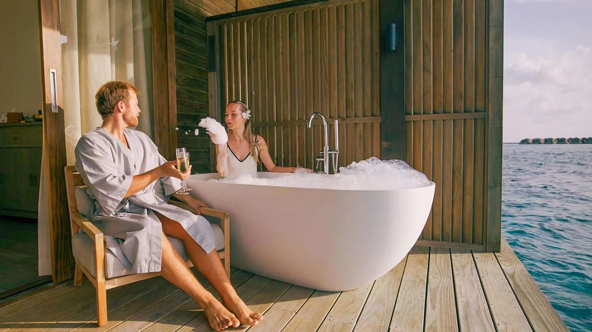 Nala Maldives by Jawakara, spa, Lhaviyani Atoll, Maldives, Overwater Spa couples-bath suite with a freestanding oval white tub on the deck, a guest in a bubble bath, a partner in a robe lifting a champagne flute beside the tub, and the wood-slat overwater wall opening to the lagoon.
