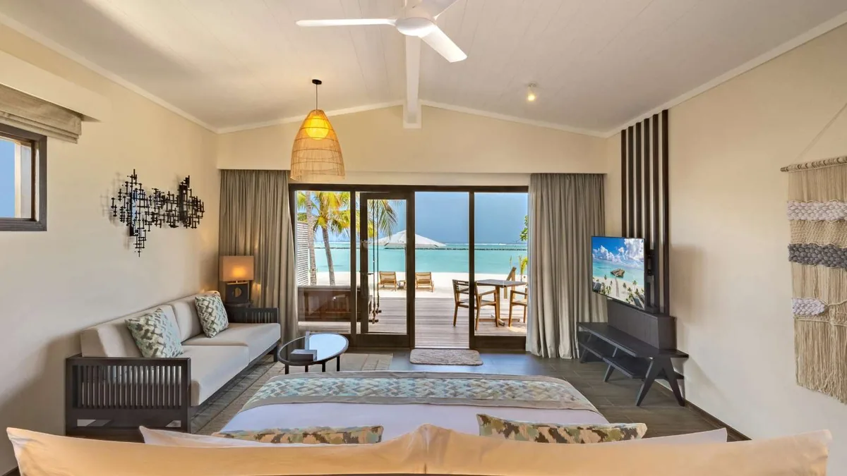 Nala Maldives by Jawakara, villa beach, Lhaviyani Atoll, Maldives, Romantic Beach Villa interior with a vaulted white ceiling and ceiling fan, a basket pendant light over the bed, an abstract metal wall installation, woven macrame wall hanging, and opened sliding doors framing the beach deck and lagoon.
