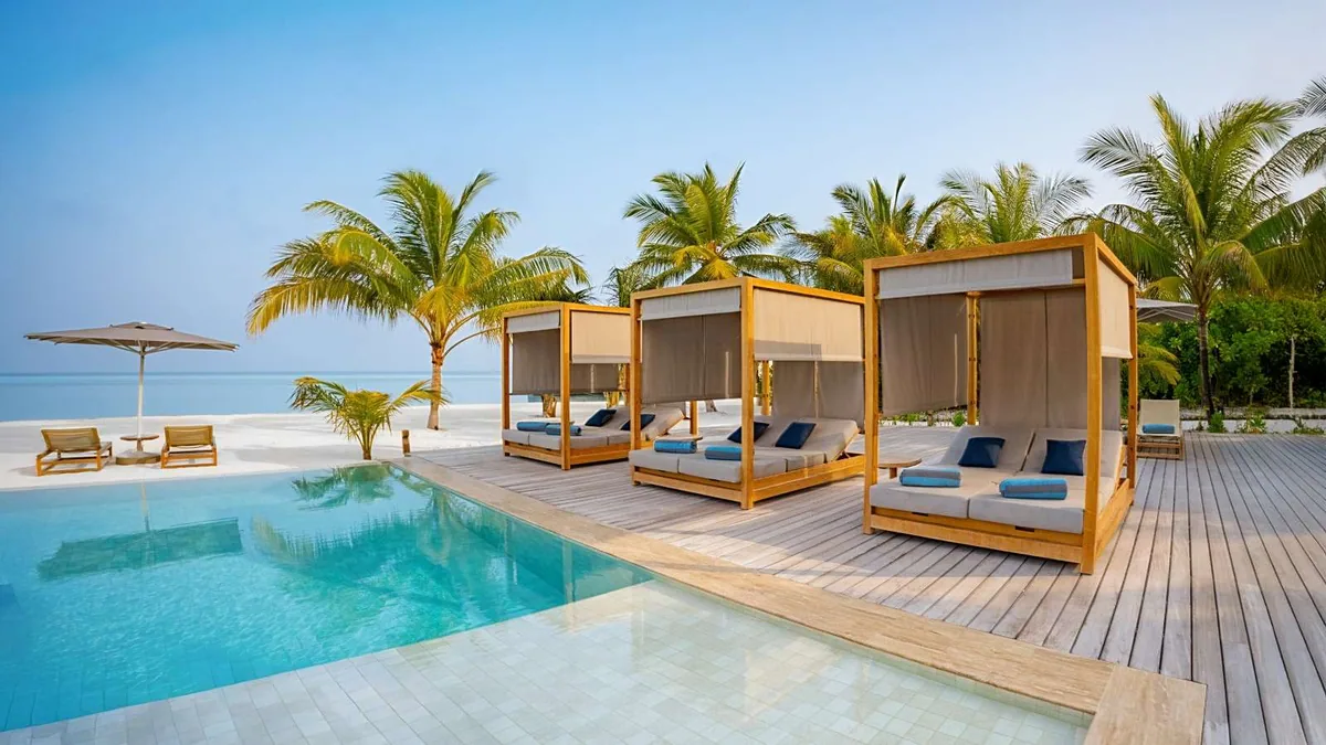 Nala Maldives by Jawakara, setting, Lhaviyani Atoll, Maldives, Pool deck with three teak-frame cabanas along the infinity-edge pool, grey cushions with blue accent pillows, palm-fringed beach line and the lagoon stretching beyond.