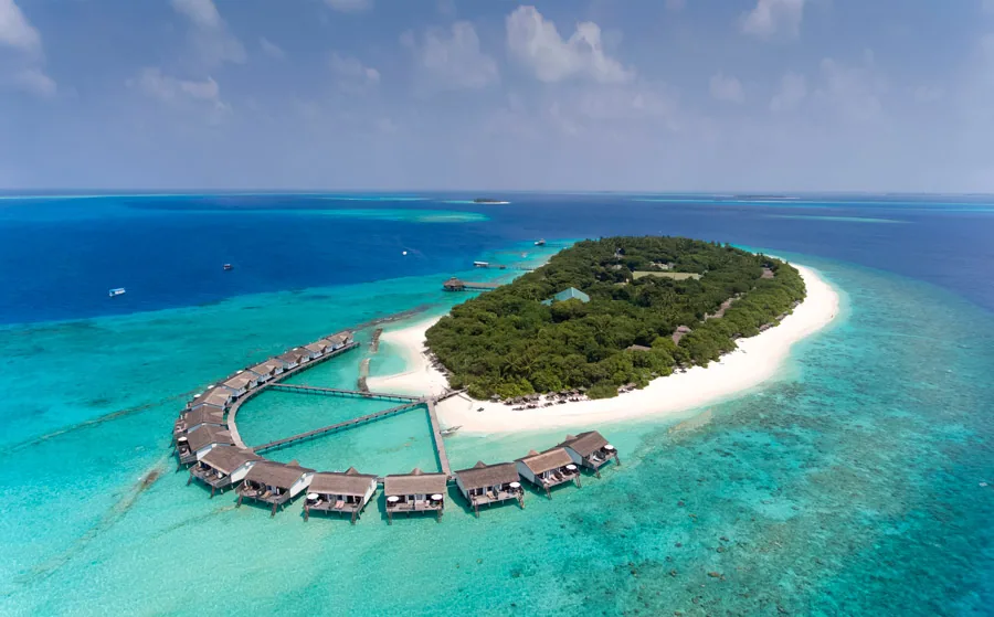 NH Collection Maldives Reethi Resort, aerial, Baa Atoll, Maldives, Overwater villa U-cluster from the lower drone angle, the curved arm of thatched-roof water villas reaching off the west jetty with the connecting triangular boardwalk, the central island bay with white sand and dense palm canopy behind.