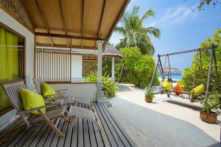 NH Collection Maldives Reethi Resort, villa beach, Baa Atoll, Maldives, Deluxe Beach Villa covered deck, twin teak sun-loungers with lime-green cushions on the timber decking, a twin hemp-rope swing chair with orange-and-green pillows beside the planted palm path leading out to the white-sand beach and lagoon.