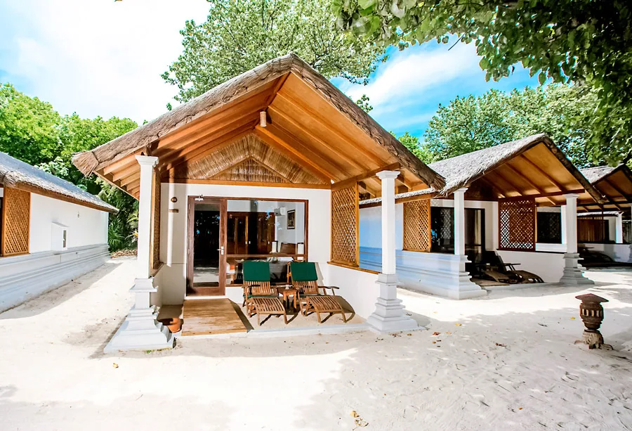 NH Collection Maldives Reethi Resort, villa beach, Baa Atoll, Maldives, Reethi Villa exterior pair, the traditional Maldivian-style thatched-roof villa structures with white classical columns and woven-cane wall panels, twin green-cushioned wood sun-loungers on the covered deck and the dense beach-vegetation canopy framing the white-sand frontage.