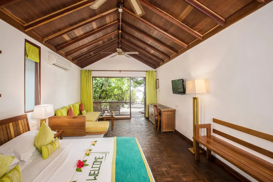 NH Collection Maldives Reethi Resort, villa beach, Baa Atoll, Maldives, Deluxe Beach Villa interior, the warm-wood gabled timber ceiling above the king bed with welcome-flower lettering on the white linen, the extensive seating area with lime-green cushioned banquette bench and the open door to the covered deck and beach.