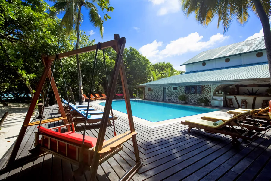 NH Collection Maldives Reethi Resort, setting, Baa Atoll, Maldives, Main resort pool deck, the rectangular swimming pool with stone-cladded back building behind, twin red-cushioned wood swing benches in the foreground on the timber decking and the cream sun-loungers lining the pool edge under the palm canopy.