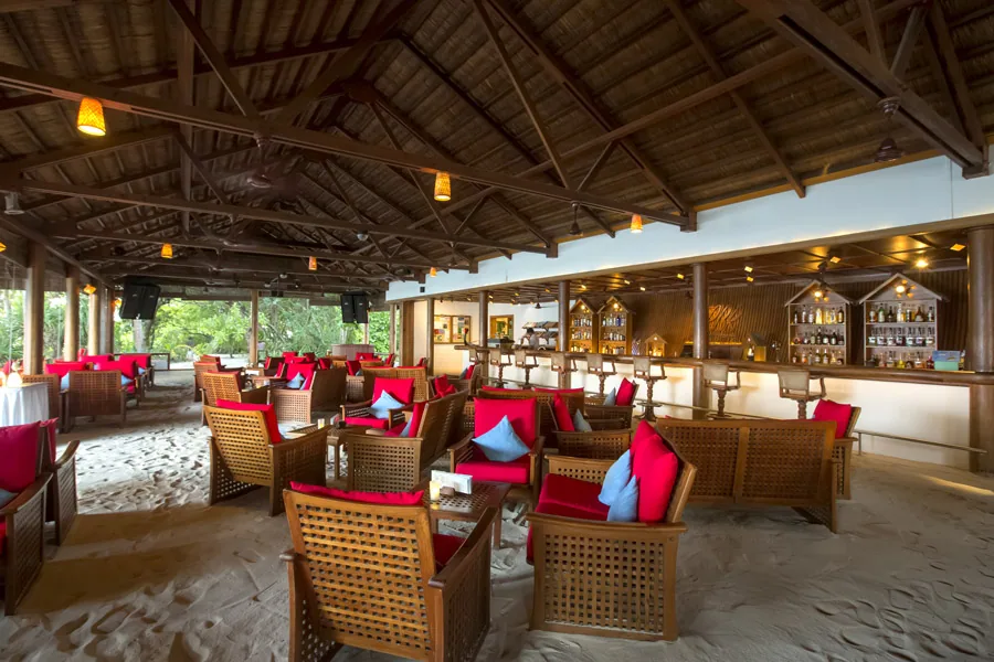 NH Collection Maldives Reethi Resort, dining, Baa Atoll, Maldives, Rasgefaanu bar interior, the open-eave thatched-frame pavilion with the red-and-blue cushioned woven-wood chairs on the soft-sand floor and the back-bar bottle wall illuminated by basket-shaded pendant lights.
