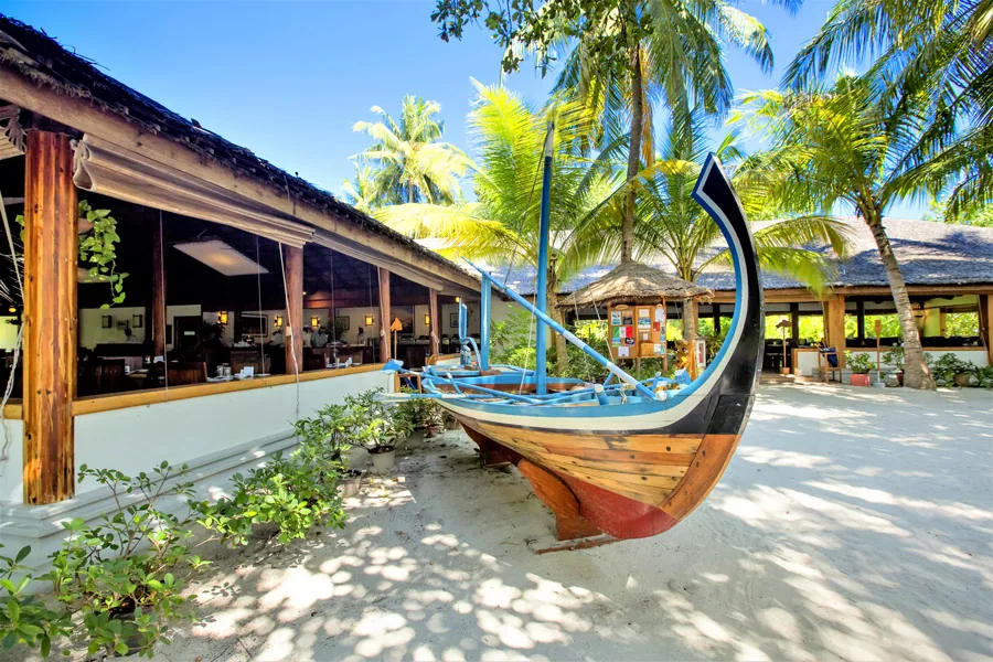 NH Collection Maldives Reethi Resort, dining, Baa Atoll, Maldives, Rehendi principal restaurant exterior, the decorative traditional Maldivian dhoni boat displayed on the white sand in front of the open-eave thatched dining pavilion, palm trees framing the entry path.