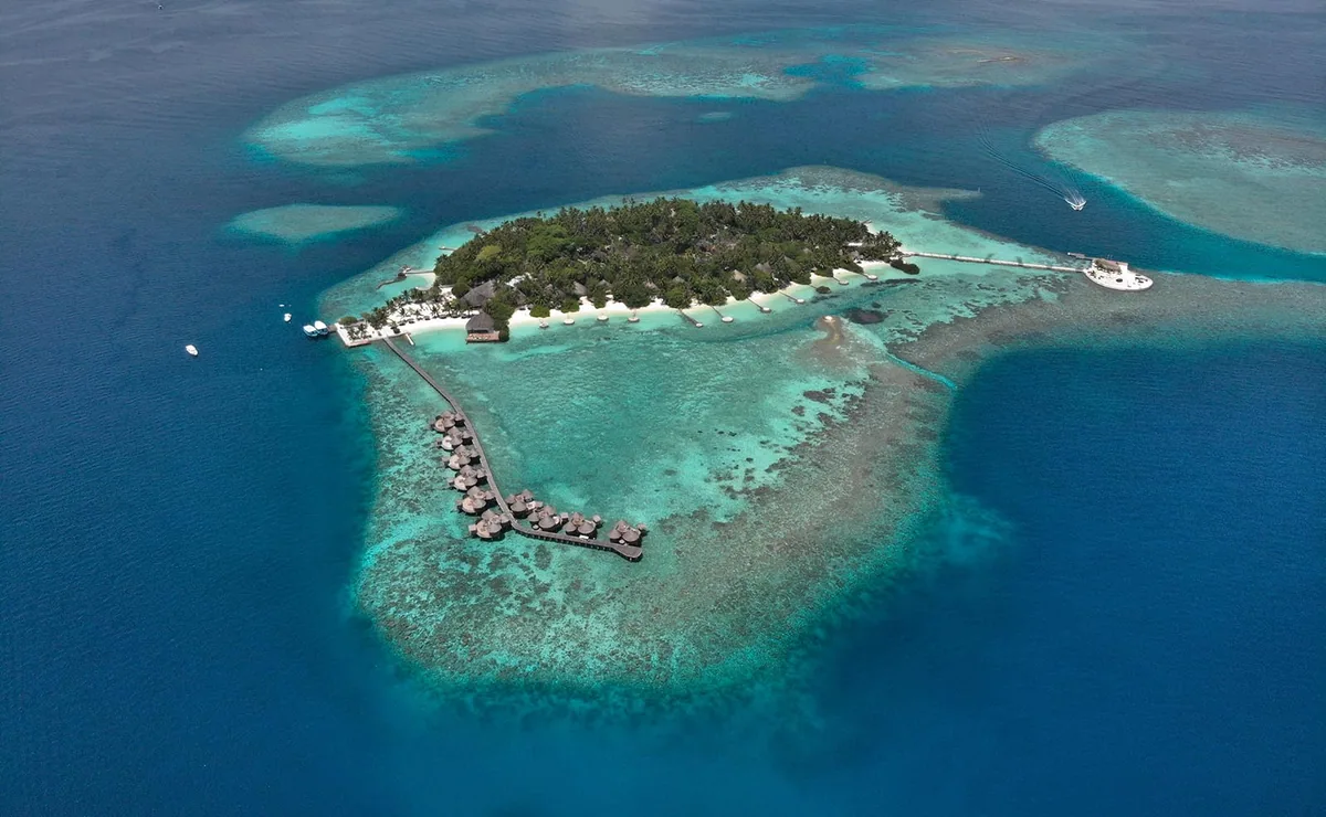 Nika Island Resort & Spa, aerial, Alif Alif Atoll (North Ari), Maldives, DJI drone aerial of Nika Island, the small palm-covered Kudafolhudhoo island with the curved hook-shaped overwater villa row (signature round conical thatched roofs) extending into the reef-protected lagoon, deep open ocean encircling.