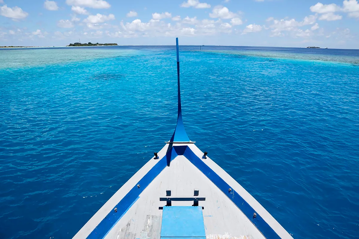 Nika Island Resort & Spa, lifestyle, Alif Alif Atoll (North Ari), Maldives, The signature Nika dhoni bow with blue-and-white painted hull cutting across the deep-blue lagoon, the elongated traditional Maldivian boat-bow ornament rising above the waterline, distant atoll islands marking the horizon.