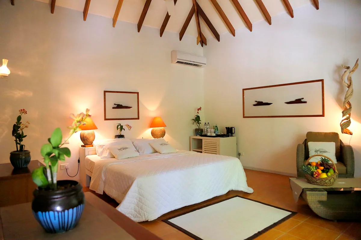 Nika Island Resort & Spa, villa beach, Alif Alif Atoll (North Ari), Maldives, Garden Room interior with white-walled cathedral pitched-rafter ceiling, king bed dressed in white linen, framed driftwood-art panels above the headboard, mango-yellow lamp shades with cone-textured bases, blue-striped pottery planters and a vintage Maldivian fishing-dhoni photograph as the room's cultural anchor.