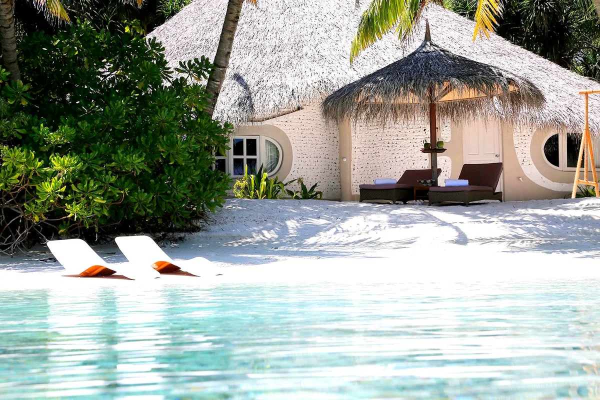 Nika Island Resort & Spa, villa beach, Alif Alif Atoll (North Ari), Maldives, Deluxe Beach Villa exterior with porthole-style round windows on the white coral-stone walls (Nika-signature ship-window architecture), thatched conical roof, sun-loungers on the white sand with orange cushions, palm canopy framing the entry path and turquoise lagoon at the foreground.