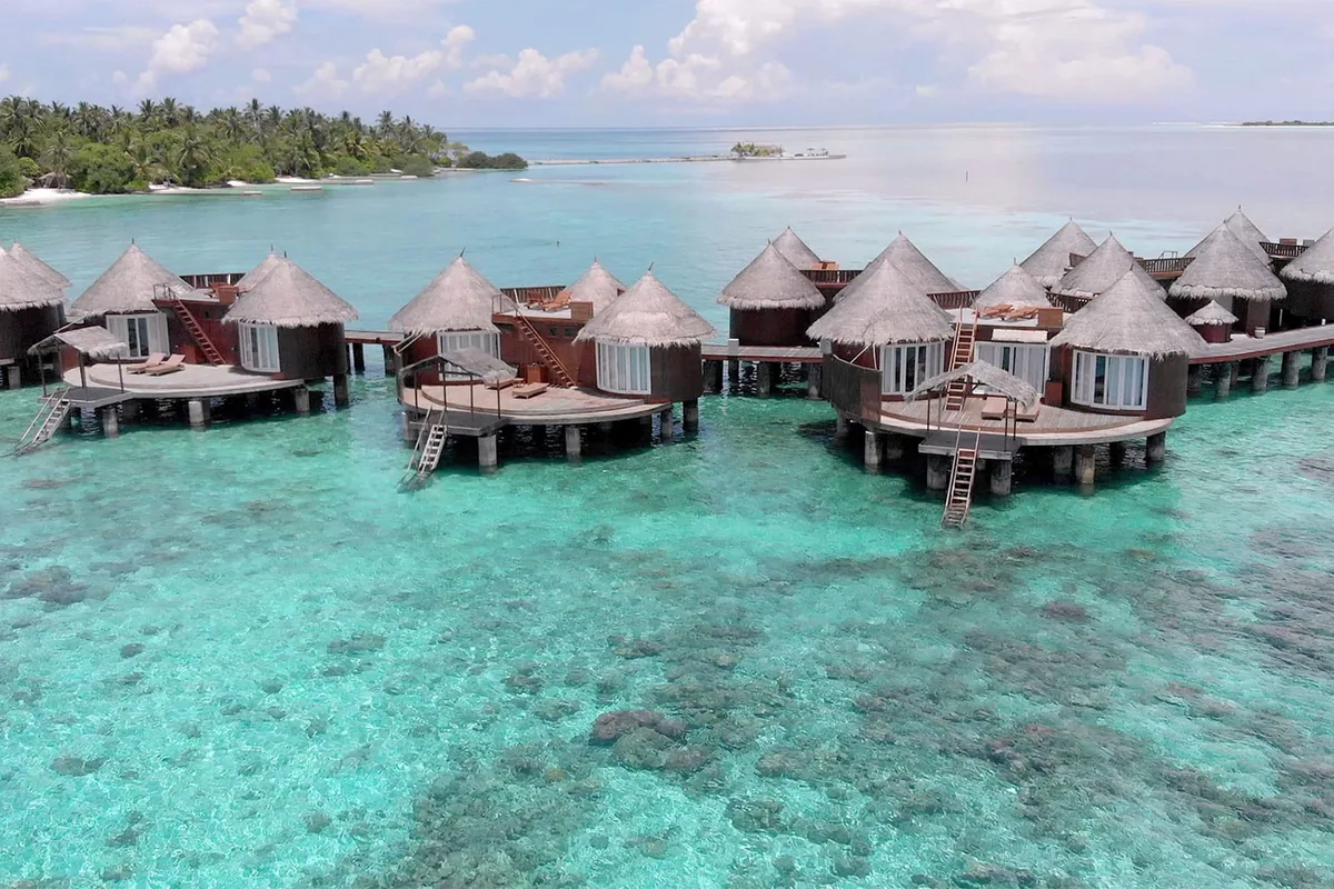 Nika Island Resort & Spa, villa overwater, Alif Alif Atoll (North Ari), Maldives, Water Villa cluster aerial showing the round conical thatched-roof OWVs (Nika's hallmark architectural form distinct from rectangular-pavilion Maldivian standard), white-walled bases over the turquoise lagoon, palm-covered main island in the background.