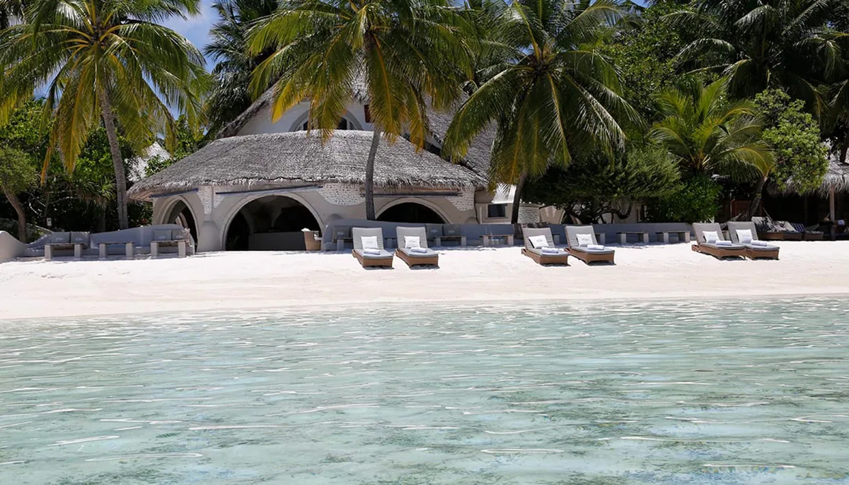 Nika Island Resort & Spa, villa beach, Alif Alif Atoll (North Ari), Maldives, Faro Studio beachside pavilion, thatched-roof villa with three arched openings across the white coral-stone facade (traditional Maldivian vernacular signature), sun-loungers and woven daybeds lined along the white sand under the palm canopy.