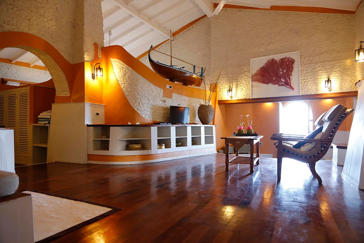 Nika Island Resort & Spa, villa suite, Alif Alif Atoll (North Ari), Maldives, Sultan Suite interior with high vaulted white-painted coral-stone walls and a bright-orange feature wall (Nika brand colour), exposed timber rafters, a model dhoni mounted on the wall, polished hardwood floor and Maldivian woven textiles framing the carved-wood reading chair.