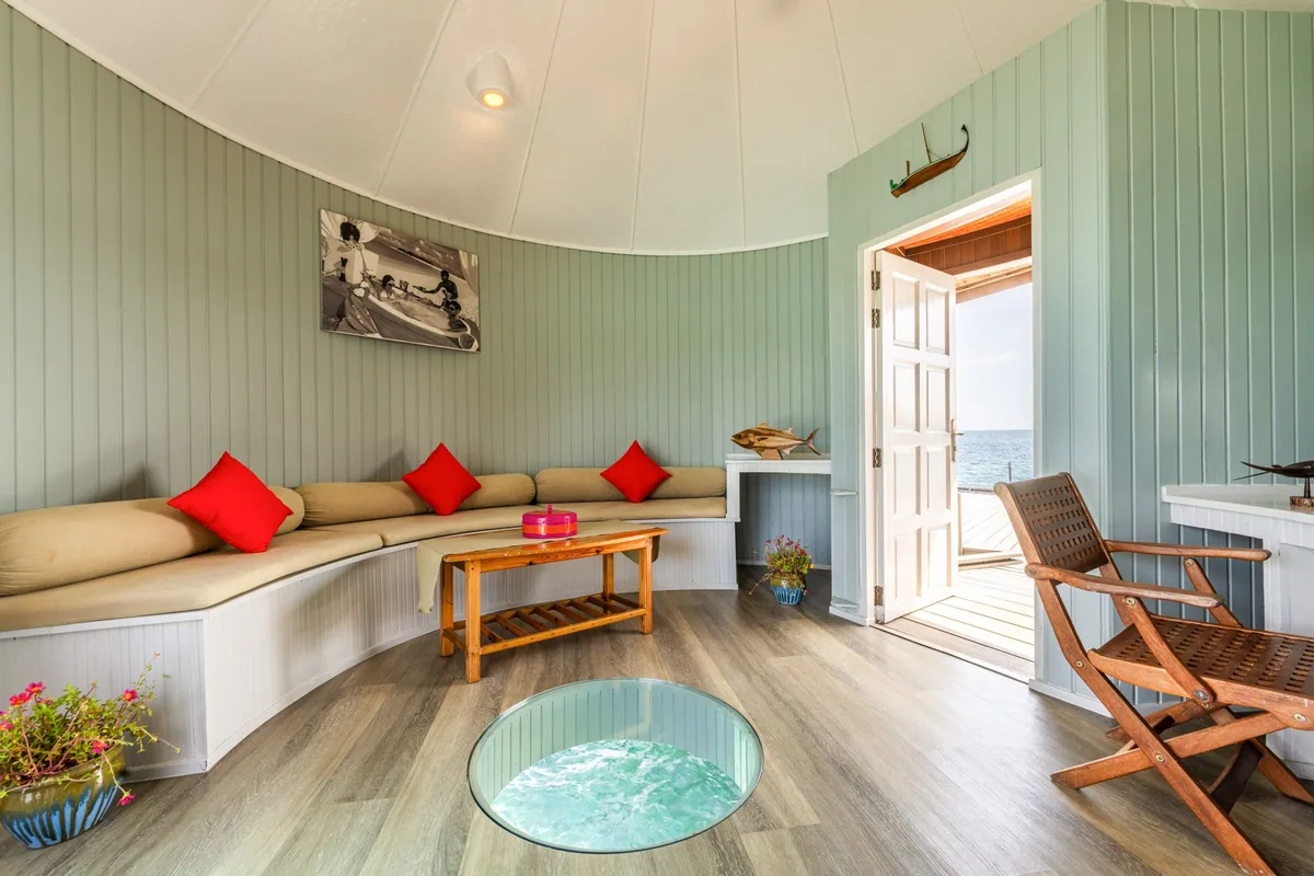 Nika Island Resort & Spa, villa overwater, Alif Alif Atoll (North Ari), Maldives, Water Villa interior with the conical-walled round room shape mirroring the exterior thatched silhouette, mint-green vertical-paneled walls, U-shaped cream banquette with red square cushions, central glass-bottom floor port revealing the lagoon water beneath, vintage Maldivian photograph and carved-fish artwork on the wall.