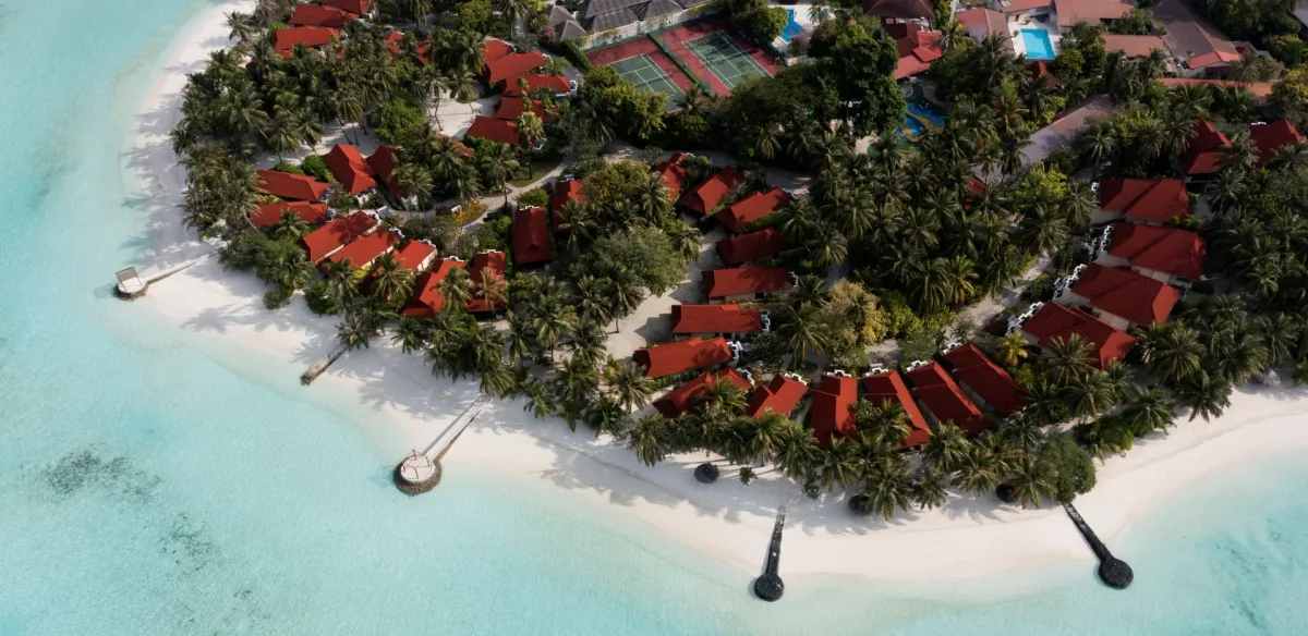 Niva Kurumba Maldives, aerial, North Malé Atoll, Maldives, Kurumba drone oblique aerial: the property's signature terracotta-tiled red-roof bungalow cluster nestled in the coconut-palm canopy along the southern Vihamanaafushi shoreline, tennis court and pool at the upper edge, white-sand beach and turquoise lagoon framing the perimeter, 4 km east of Velana International.