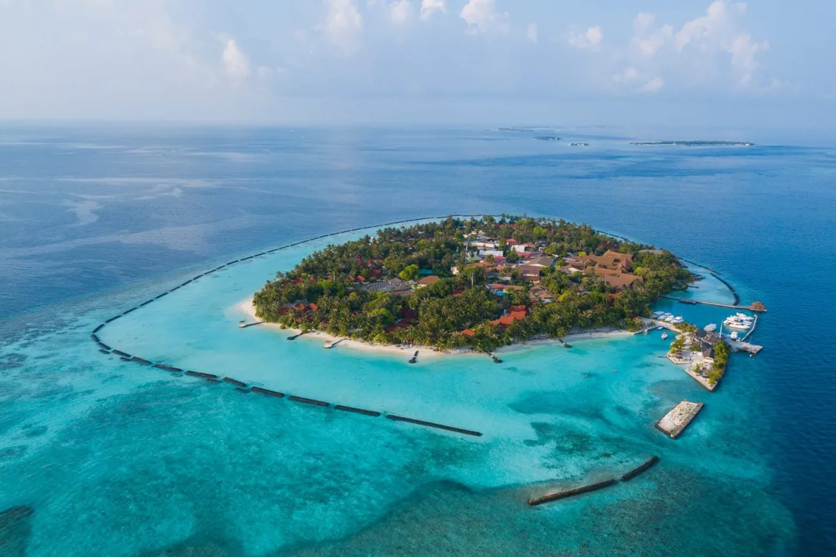 Niva Kurumba Maldives, aerial, North Malé Atoll, Maldives, Wide bird's-eye Vihamanaafushi aerial: the full oval-shape palm-canopied island with the distinctive red-roof bungalow cluster visible through the trees, surrounding breakwater protecting the shallow lagoon, the marina with boats at the right edge, neighbouring small sandbar visible at the far edge.