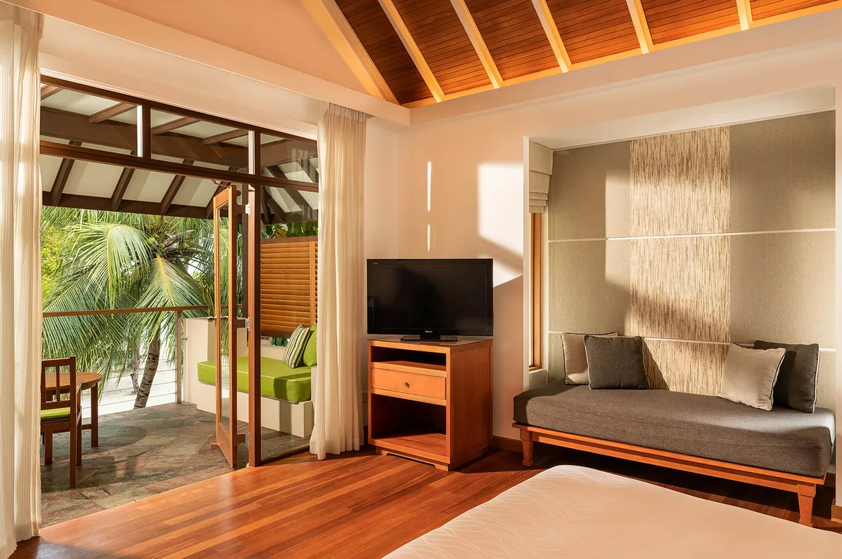 Niva Kurumba Maldives, villa beach, North Malé Atoll, Maldives, Superior Bungalow interior: vaulted wooden-beam ceiling, TV on light-wood cabinet, neutral daybed positioned against a textile-panelled feature wall, sliding glass door opening onto a private terrace with a green cushion bench and tropical palm view beyond.