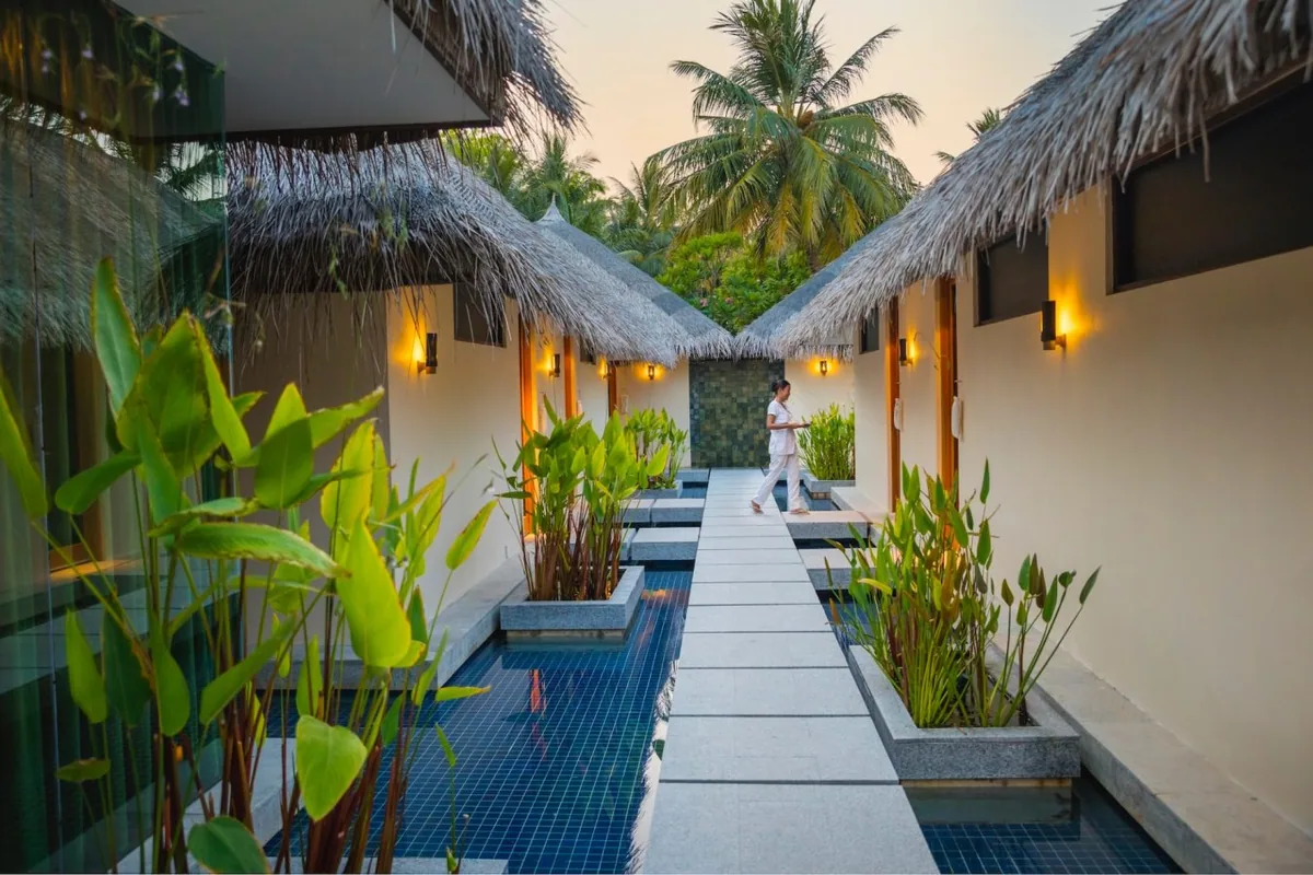 Niva Kurumba Maldives, spa, North Malé Atoll, Maldives, Veli Spa entrance walkway: twin thatched-roof spa pavilions facing each other with a stone walkway between them flanked by twin water-channels with deep-blue mosaic tile and tropical heliconia plants, terracotta stepping-stones in the channels, wall sconces, a guest in a white robe walking down the walkway at dusk.