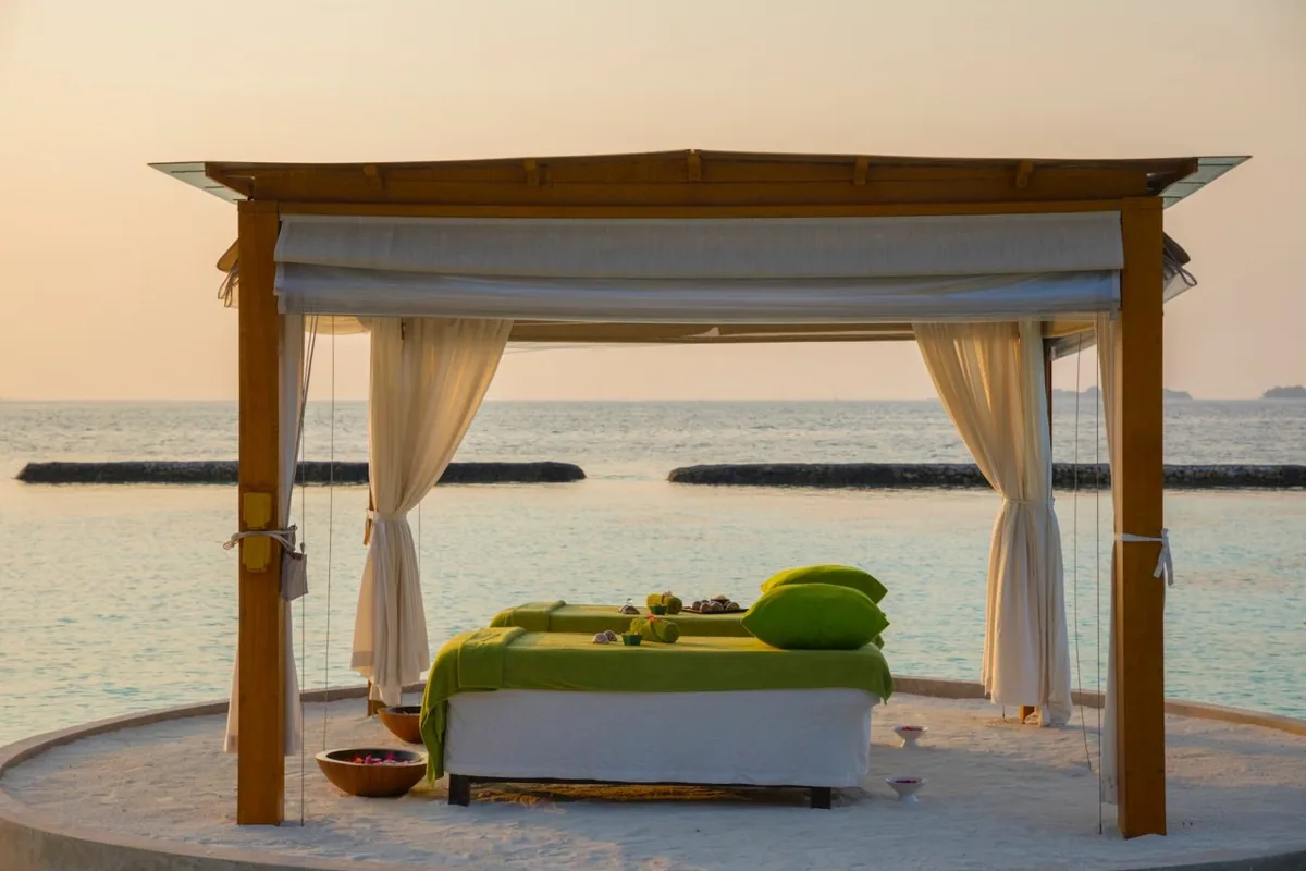 Niva Kurumba Maldives, spa, North Malé Atoll, Maldives, Outdoor beach-edge spa gazebo at sunset: open-sided timber-pillared gazebo with a pitched roof, twin massage beds dressed in lime-green linens, gauzy white curtain drapes tied at the corner posts, candle bowls and ritual prep arranged on the sand, the calm sea with breakwater rocks visible behind, a golden-orange sunset sky.