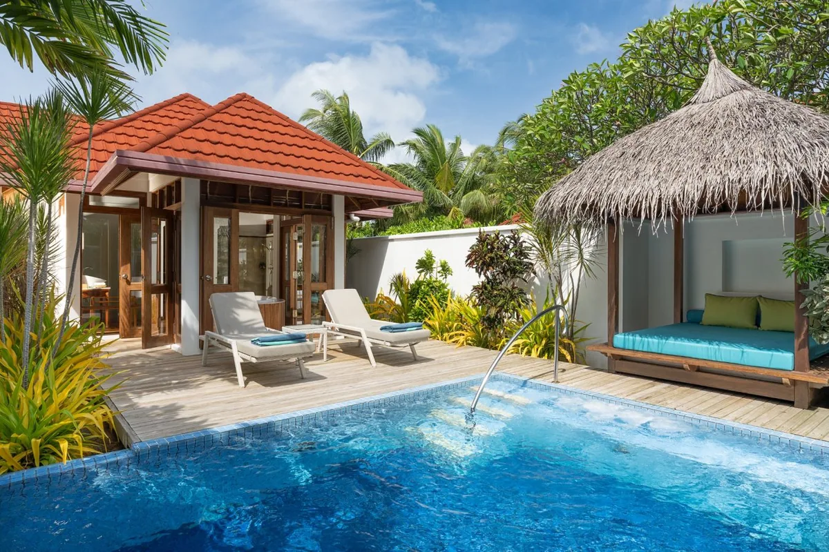 Niva Kurumba Maldives, villa beach, North Malé Atoll, Maldives, Two-Bedroom Kurumba Residence exterior with private pool: signature red-tile pitched-roof bungalow flanked by twin sun loungers on a timber deck, an attached thatched-roof beach cabana with cushioned daybed in green, a private rectangular plunge pool with water-spouts cascading down the edge, tropical garden plantings framing the courtyard.