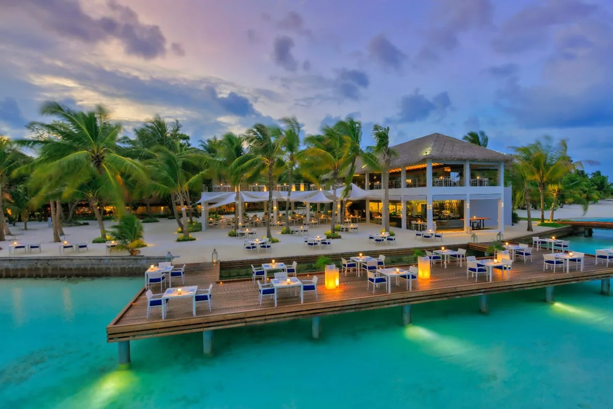 Niva Kurumba Maldives, dining, North Malé Atoll, Maldives, Thila restaurant at twilight: large two-storey thatched-roof open pavilion plus an over-water timber deck extending into the lagoon with dining tables set, candle lanterns at each table, white sail awnings on the beach-side terrace, illuminated underwater lights creating an aqua glow in the lagoon, a dramatic peach-purple twilight sky, dense palm canopy framing the back.