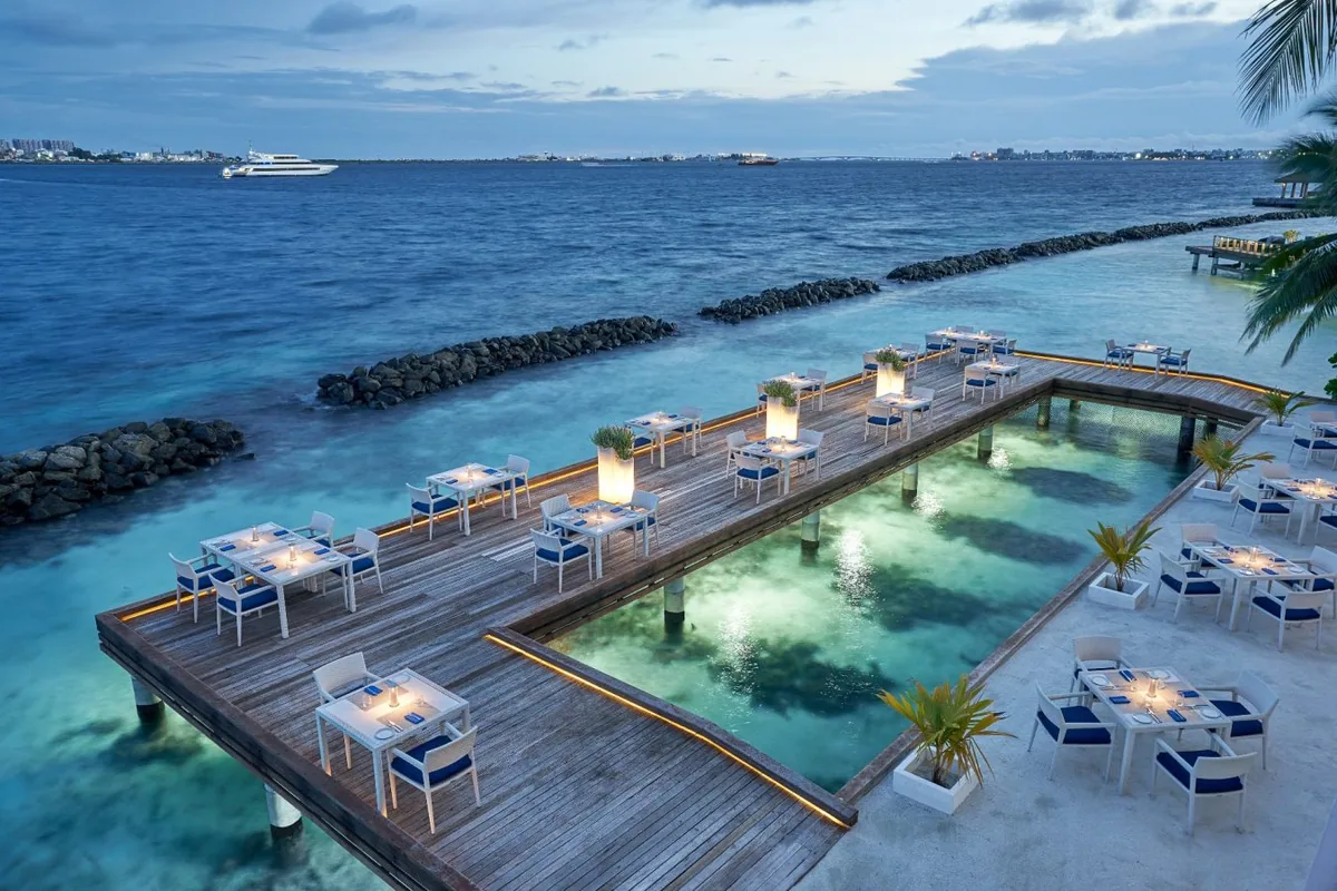Niva Kurumba Maldives, dining, North Malé Atoll, Maldives, Thila over-water deck close-up: U-shaped timber deck with dining tables (white-and-blue cushioned chairs) wrapping around a central illuminated swimming pool with glowing aqua light, candle lanterns at each table, the Velana airport lights and ships visible in the far distance, breakwater rocks framing the lagoon edge.