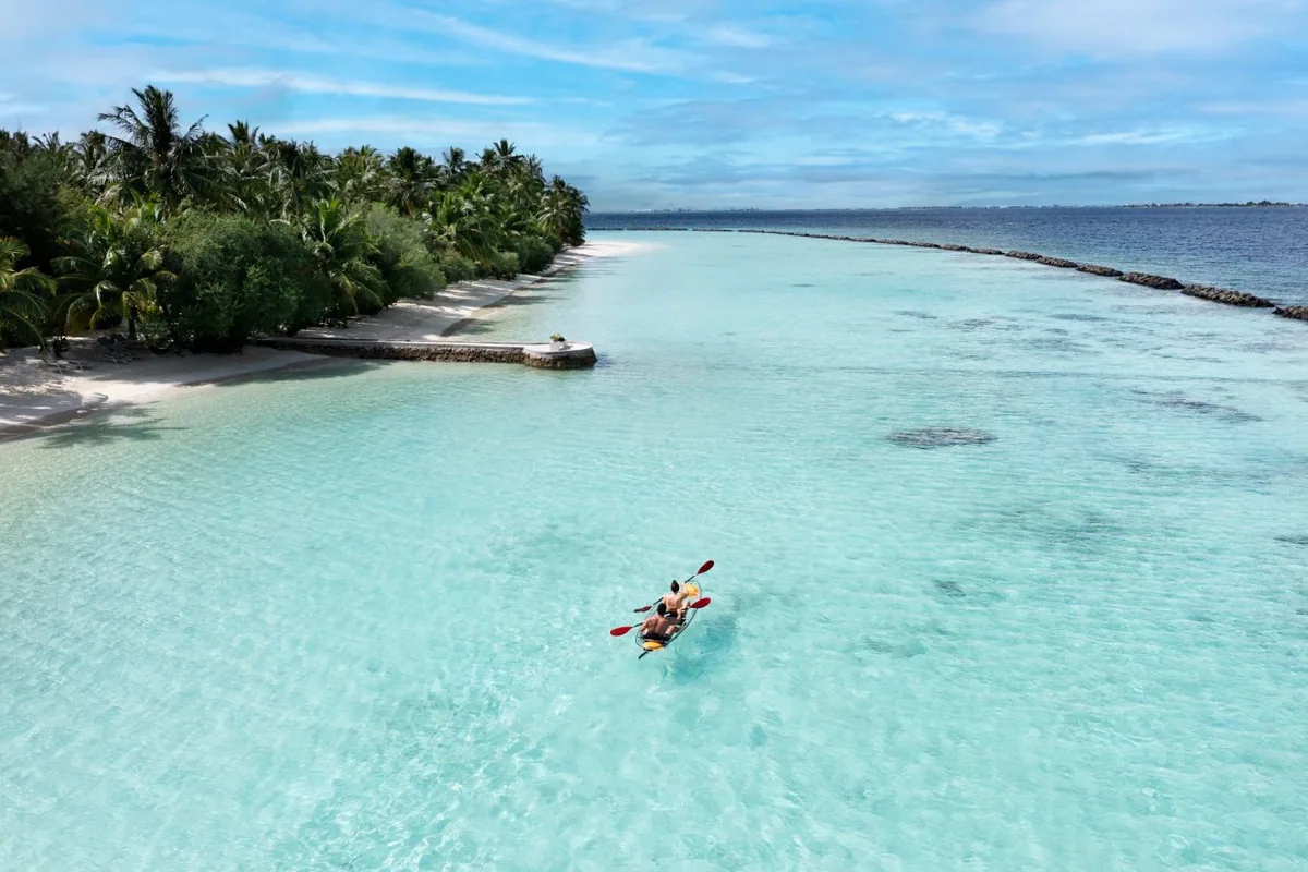 Niva Kurumba Maldives, activities, North Malé Atoll, Maldives, Lagoon kayaking aerial: a yellow tandem kayak with two paddlers gliding across the crystal-clear shallow turquoise lagoon between the palm-canopied Kurumba shore and the signature breakwater rocks defining the property's protected lagoon, the watersports programme captured in property-signature framing.