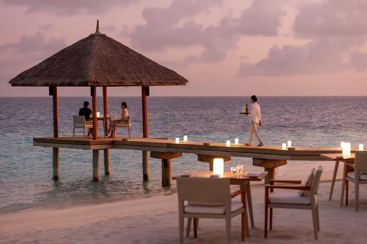 Niva Velassaru Maldives, dining, South Malé Atoll, Maldives, Sunset private dining at the overwater cabana: thatched-cone pavilion at the jetty end with a couple seated at a candle-lit table, server on the boardwalk approach with a tray, candle lanterns lining the deck rail, beach-set dining tables in the foreground, post-sunset peach sky over the lagoon.