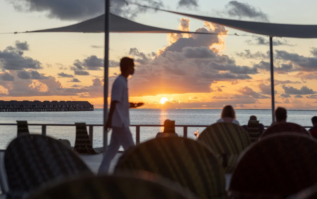 Niva Velassaru Maldives, dining, South Malé Atoll, Maldives, Chill Bar deck at sunset: silhouetted server crossing the deck with the sun dropping to the horizon, overwater villa row visible at the left edge, striped lounge seats arranged around the rail, white sailshade canopy stretched overhead.