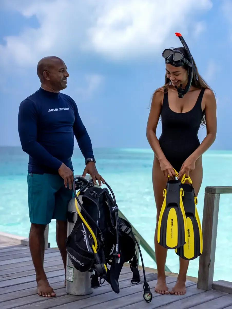 Niva Velassaru Maldives, activity, South Malé Atoll, Maldives, Aqua Sport dive centre on the jetty: instructor in the branded Aqua Sport blue rashguard holding scuba tank and BCD, guest with snorkel mask and yellow Mares fins, the turquoise lagoon and clouded sky beyond.